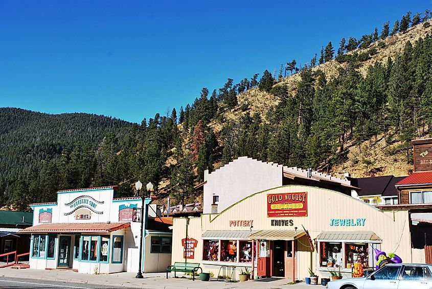 A small mountain town scene with colorful shops under a clear blue sky. A pine-covered hillside forms the backdrop, conveying a quaint and peaceful atmosphere.