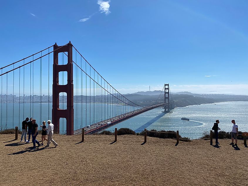 People walk on a dirt path overlooking the Golden Gate Bridge and San Francisco Bay. The sky is clear, conveying a bright, sunny day.
