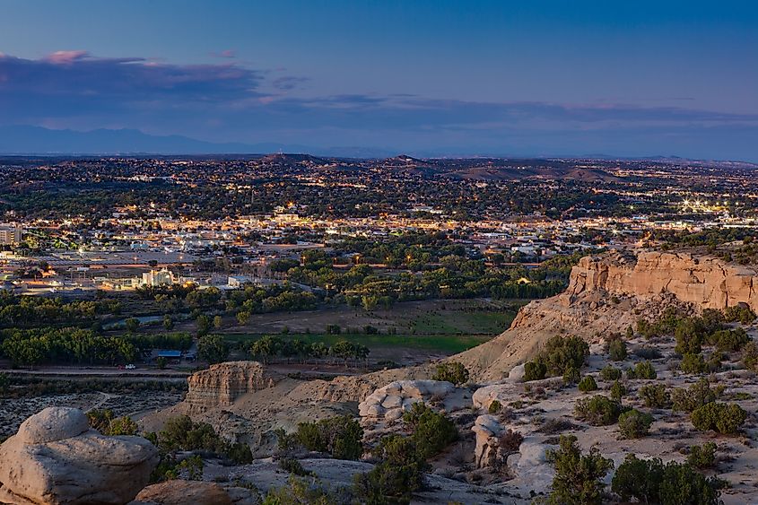 Aerial view of a city at dusk with glowing lights, surrounded by rocky cliffs and greenery. The sky is a blend of blue and purple hues, creating a serene ambiance.