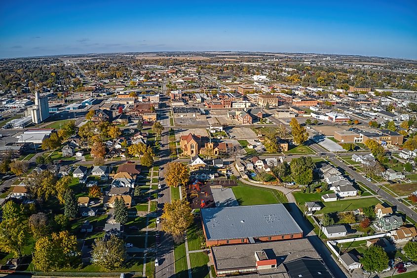 Aerial view of a small town with tree-lined streets and scattered houses. The town includes residential areas, churches, and a distant field under a clear blue sky. The atmosphere conveys a peaceful, community-focused environment.