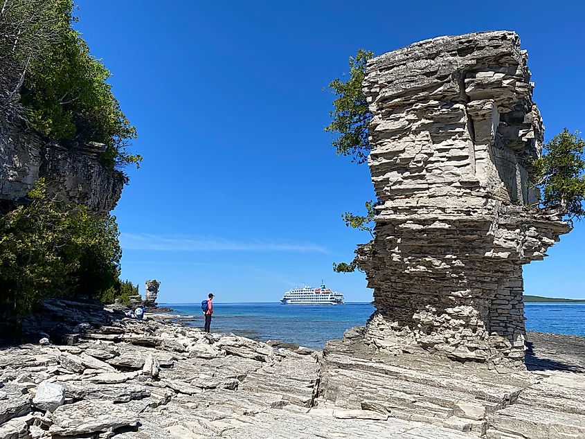 A woman in a pink jacket stands beside a limestone tower as a cruise ship passes by in the distance.