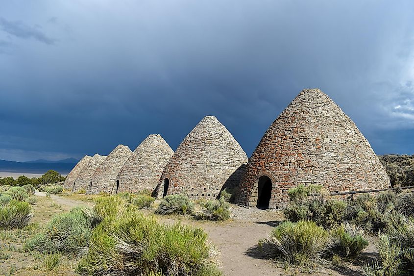  Ely, Nevada: An angled, sunny view of the six beehives at Ward Charcoal Ovens State Historic Park, set against dark, cloudy storm skies in the background.