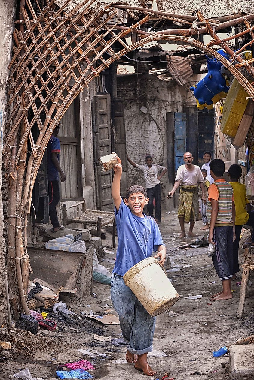 A young boy holding a bucket, joyfully running through a crowded alley in Yemen, with other residents looking on. Image Credit Rod Waddington via Wikimedia.