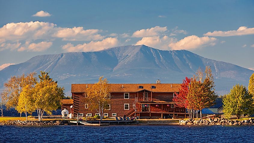 Lodge by a lake with colorful autumn trees, mountains in the background, and blue sky with scattered clouds, creating a serene and picturesque scene.