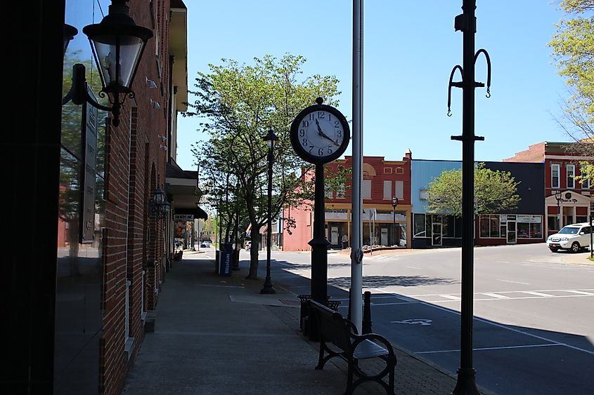 A quiet street scene in downtown Glasgow, Kentucky, showing a classic black street clock and lampposts along the sidewalk beside a brick building. Across the street, colorful historic storefronts line the opposite side, with a few trees and a parked car in the background, creating a peaceful small-town atmosphere.