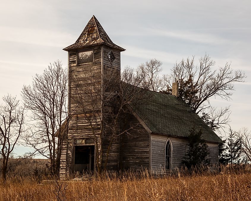 An abandoned wooden church with a tall steeple, surrounded by leafless trees, set in a field of dry grass under a cloudy sky, evokes a nostalgic mood.