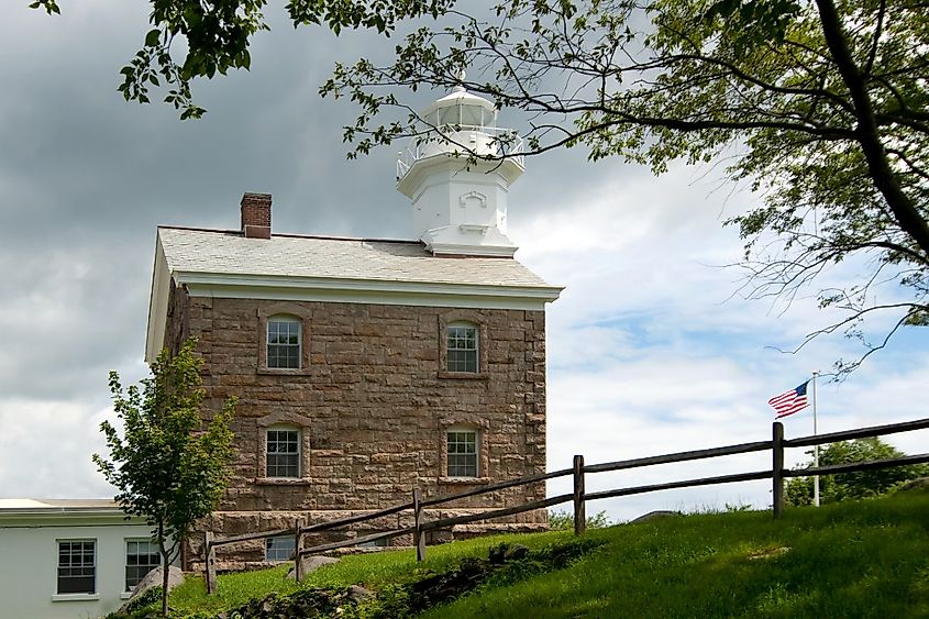 Great Captain Island lighthouse with sun shining between storm clouds. It is a favorite Connecticut attraction. American flag waving.