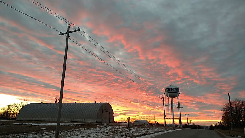 A vibrant sunset over West Point, Nebraska, with warm hues illuminating the sky and casting a glow over the town and surrounding fields.