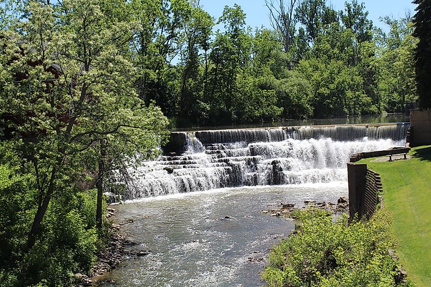 Waterfall on Honeoye Creek. Honeoye Falls, New York. Grass seating area with a bench overlooking the falls. Tree lined with blue sky in the background.