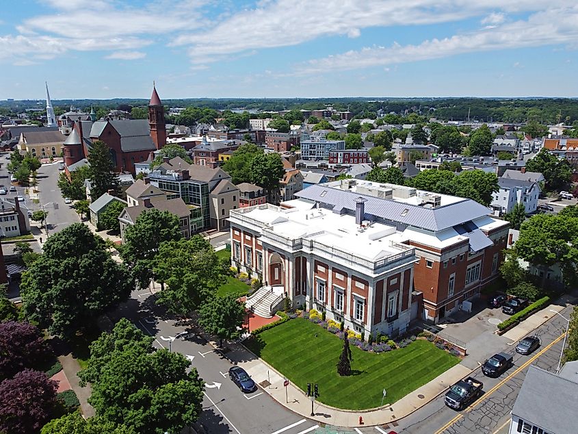 Aerial view of Beverly Public Library at 32 Essex Street, with Cabot Street and the historic city center of Beverly, Massachusetts, visible in the background. The library is surrounded by tree-lined streets and nearby buildings.