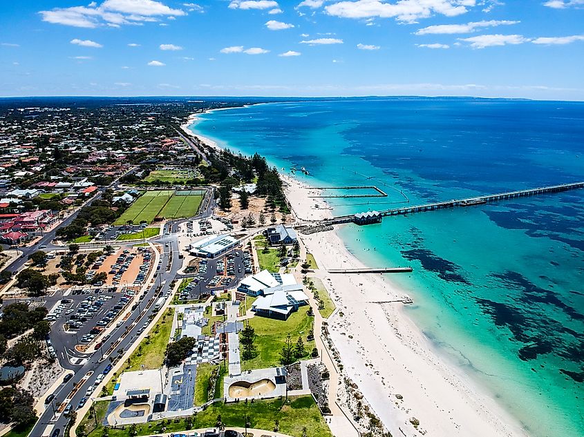 An aerial view of Busselton Jetty in Western Australia reveals the iconic wooden pier extending over the turquoise waters, with the shoreline and town of Busselton visible in the background.