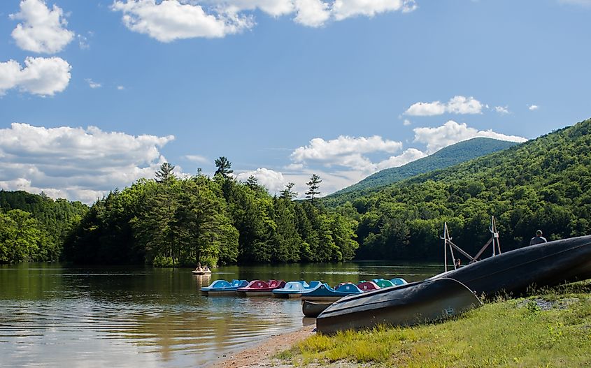 Row of paddle boats and canoes by the shore of Emerald Lake at Emerald Lake State Park in East Dorset, Vermont. The lake is encircled by lush green trees under a clear blue sky with a few clouds.