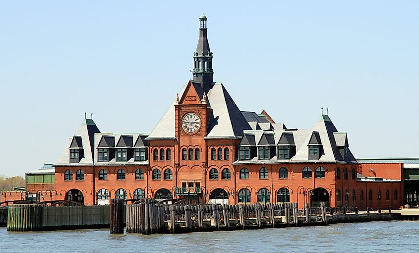 Red-brick historic train station with a green-roofed clock tower and multiple gables, set against a clear blue sky, viewed from across the water.