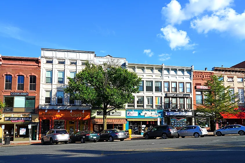 Row of historic storefront buildings with colorful facades and parked cars along a sunny main street, with a large tree in front.