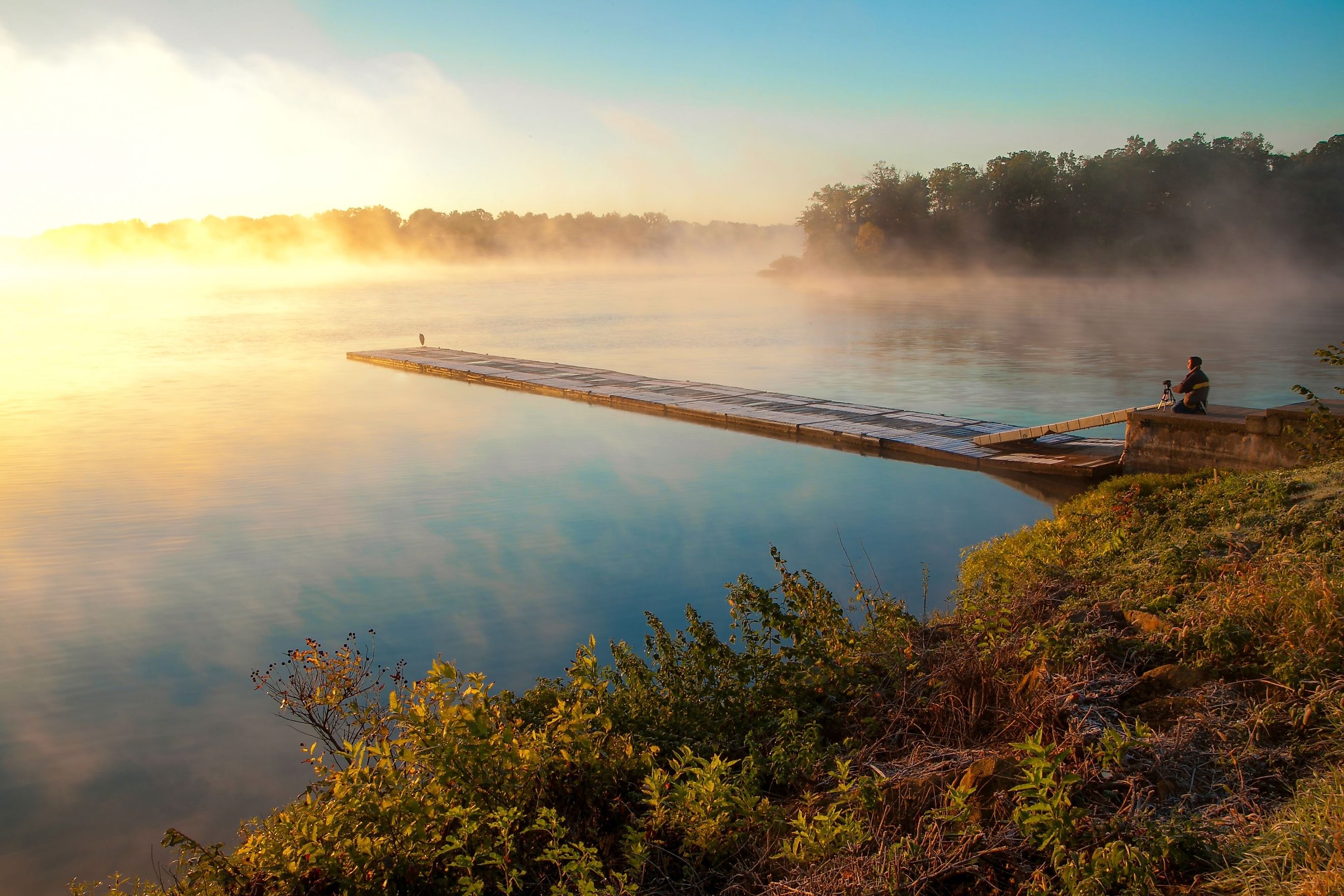 Man on a wooden boardwalk pier photographing a tranquil, fog-covered lake at sunrise