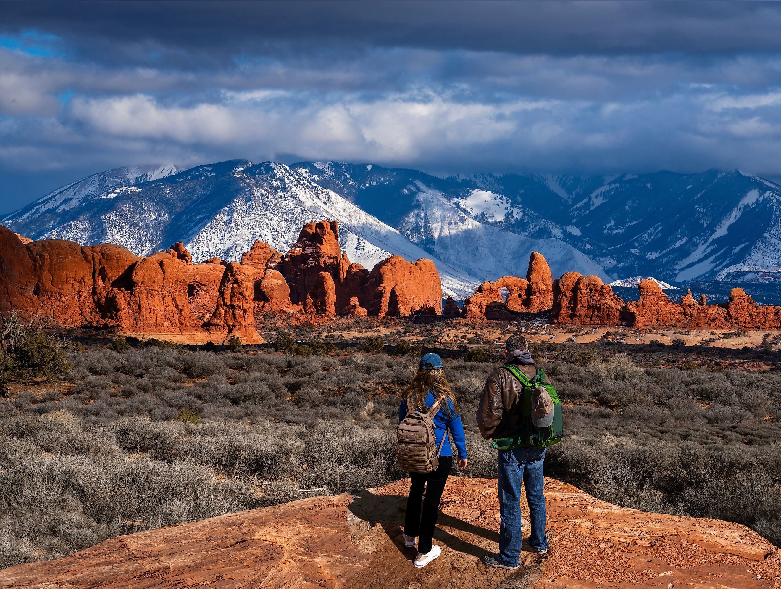 Couple enjoying a beautiful mountain view on a hiking trip in Utah, with the Windows Section of the park and snow-covered La Sal Mountains in the background. Arches National Park, Moab, Utah, USA.