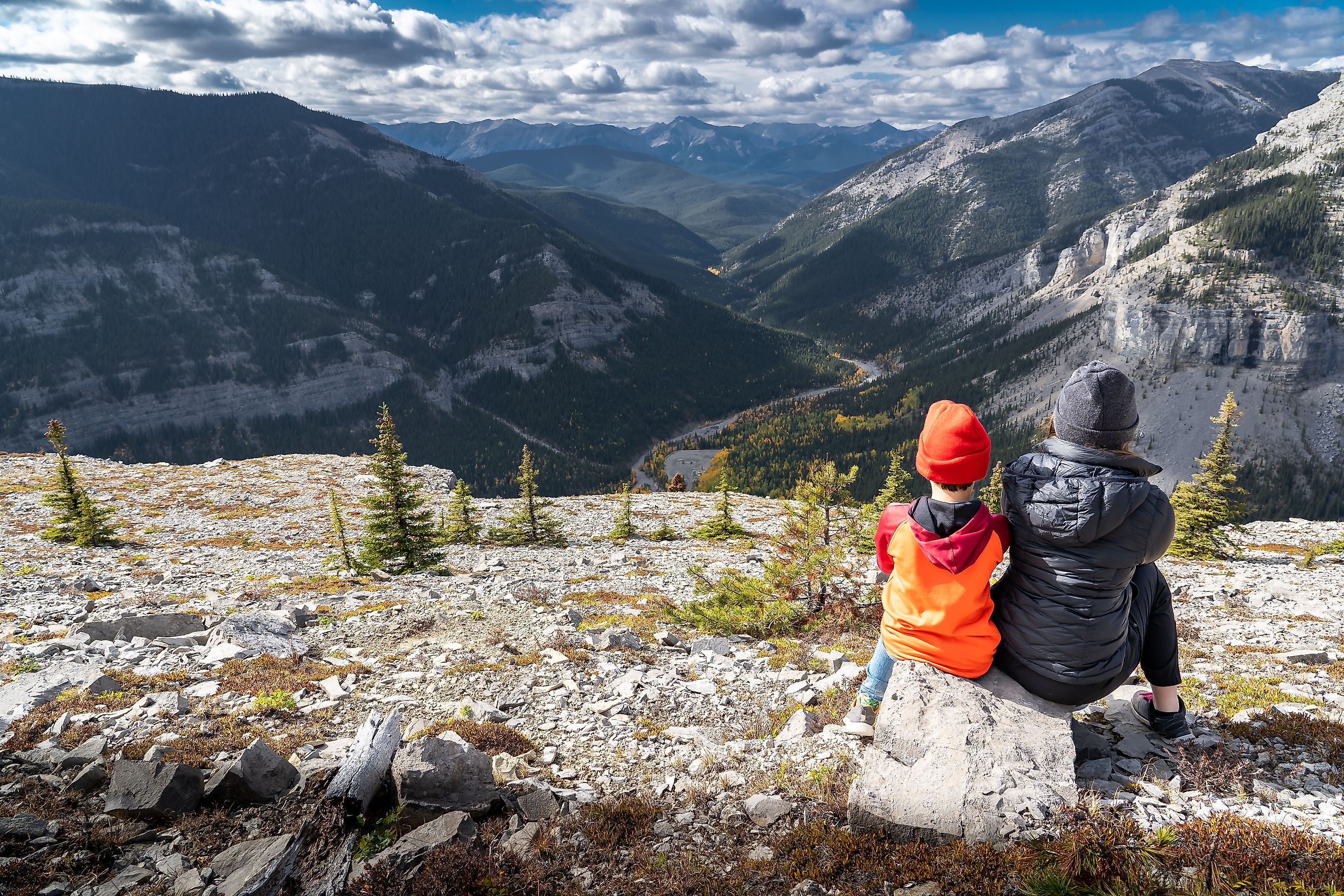 A brother and sister sitting on a rock overlooking the Canadian Rocky Mountains, with a valley of trees displaying fall colors on Moose Mountain near Bragg Creek, Alberta, Canada.