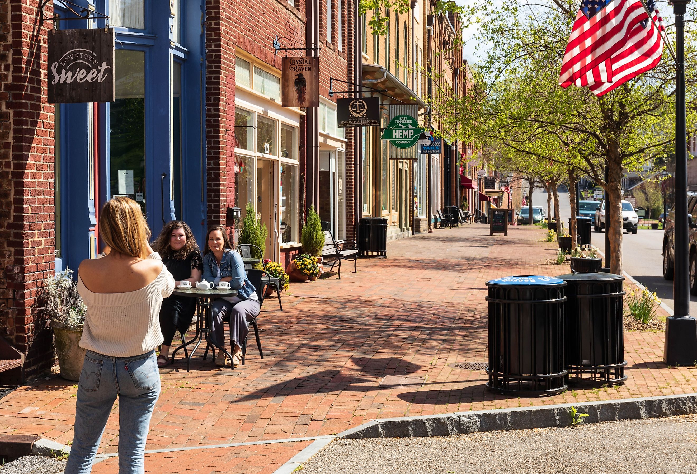 Woman with back to camera takes a picture of two friends seated at a sidewalk table, in front of the 'Downtown Sweet' coffee shop in historic Jonesborough, TN. Image credit Nolichuckyjake via Shutterstock.