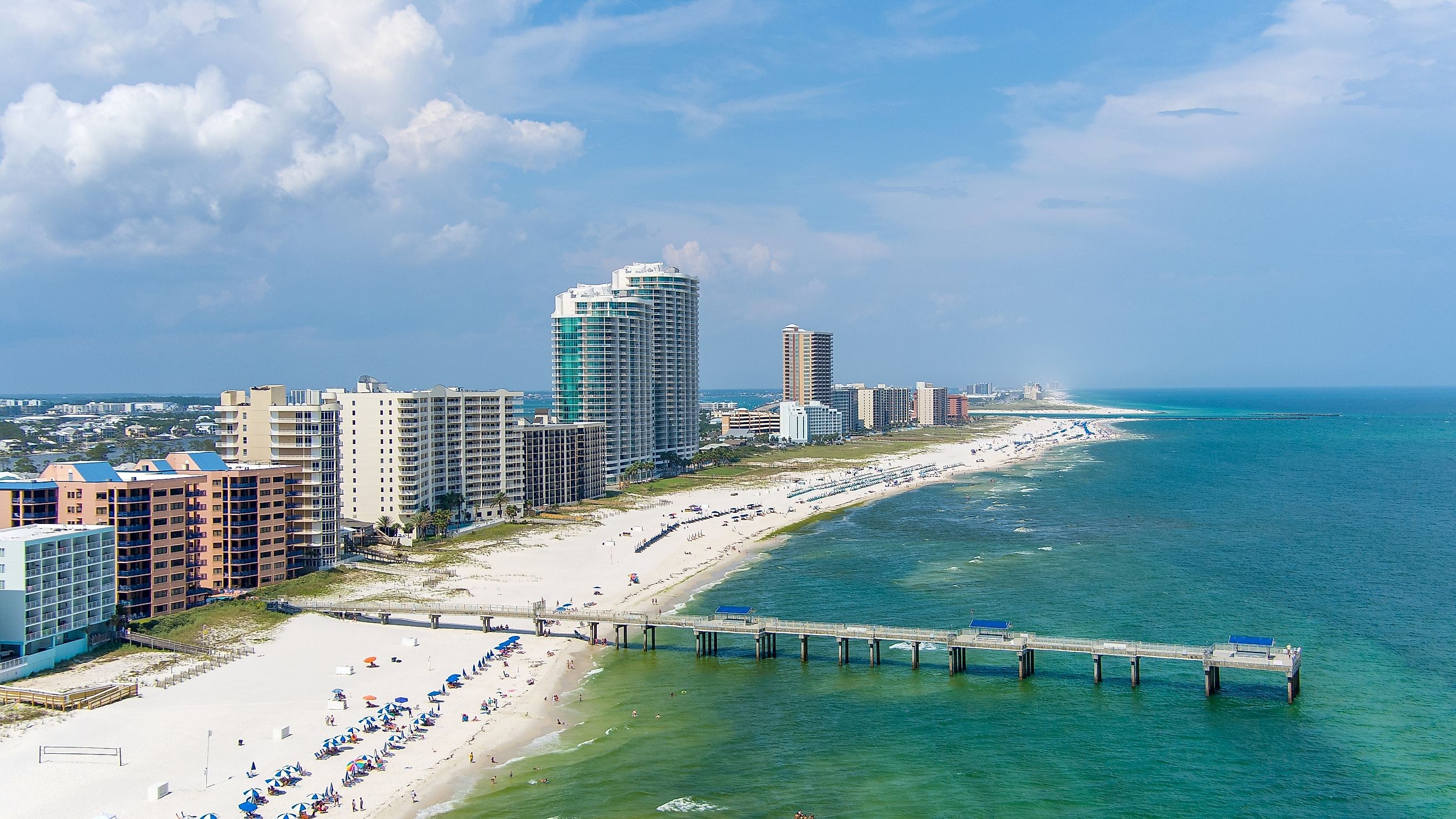 Aerial view of the beach at Orange Beach, Alabama, on a sunny day, featuring the sandy shoreline, blue ocean, and beachgoers enjoying the warm weather.