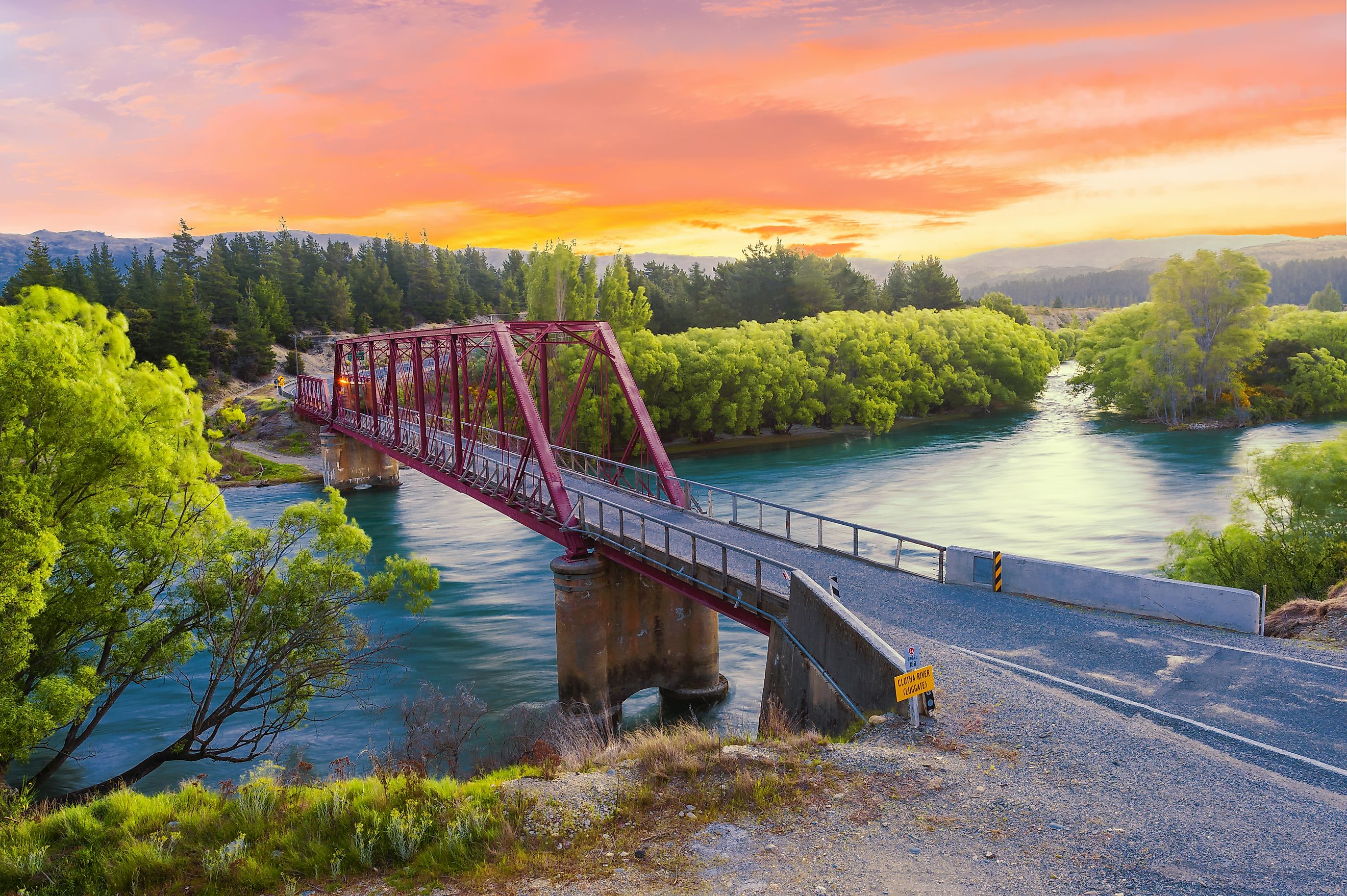 A stunning sunset illuminates the Clyde Bridge on the Clutha River, with the vibrant hues of orange and pink reflecting off the calm waters.